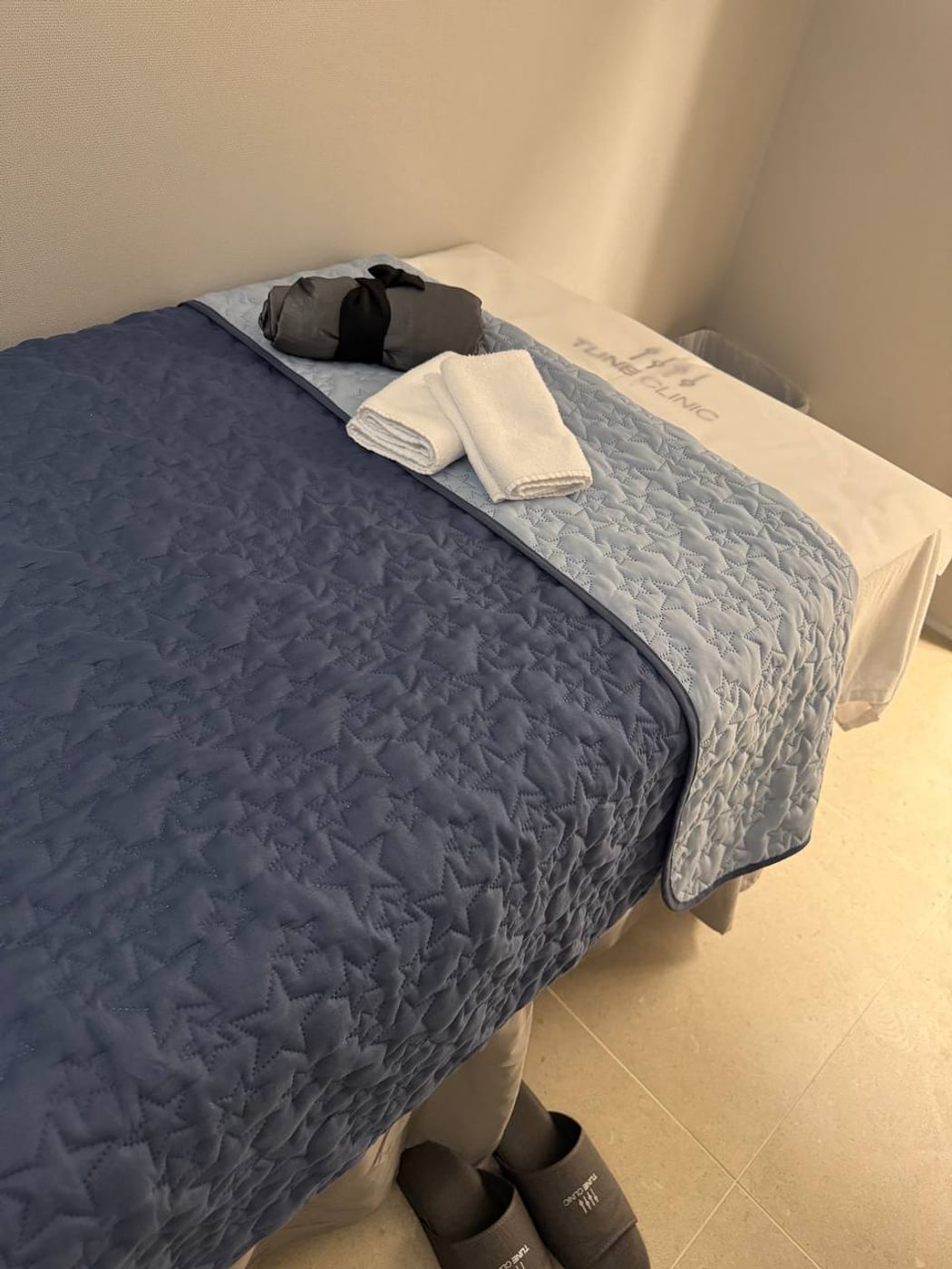 A Tune Clinic treatment bed made up with a quilted cover, folded towels and a gown on branded linen